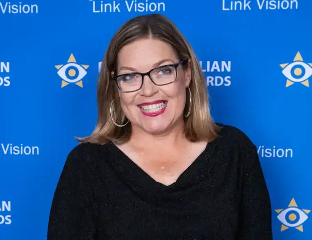 Megan is standing in front of the Blind Australian of the Year Awards and Link Vision media wall. She has a big smile and is holding her award on the red carpet.